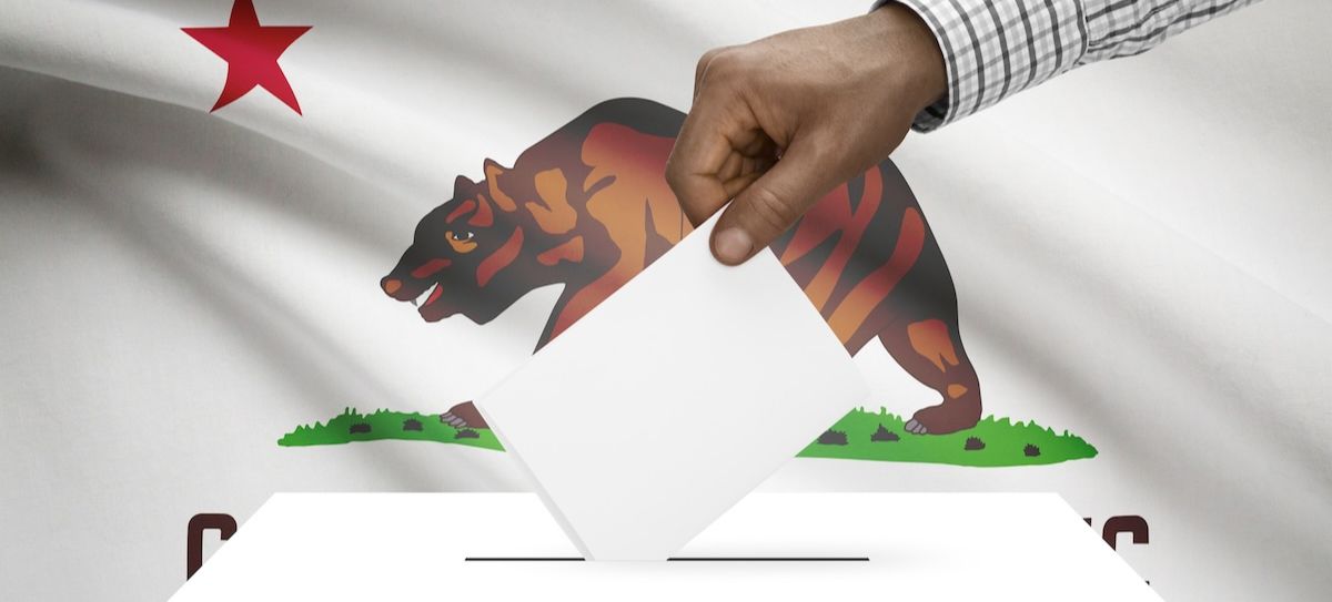 Person putting a vote into a ballot box in front of the state flag of California. The California Nations Indian Gaming Association said a new tribal-driven ballot initiative could be ready for the 2026 election cycle.