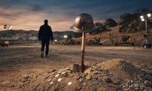A construction shovel stands upright in a pile of dirt with a hard hat on top, while a person walks away toward construction machinery at dusk.